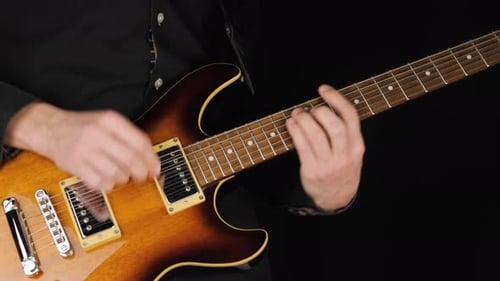 Guitarist plays a light brown six-string electric guitar with a pick against black background. 4K