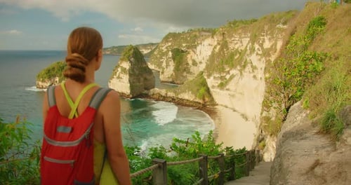 Woman traveler with backpack views ocean and rocky Diamond Beach from cliff edge on Nusa Penida