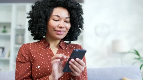 Close up. Young african american female using smartphone sitting on sofa in living room at home.