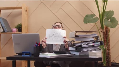 Woman office worker holds piece of paper on which it says help and shouts, laptop and stack of docum
