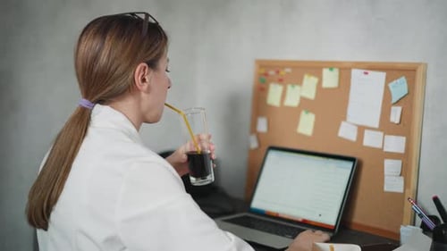 Back View of Woman Drinking Juice While Working on Laptop with Sticky Notes in Office