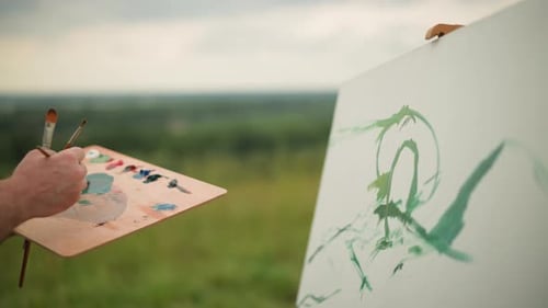 Painter Holding Palette and Brush Applying Green Paint on Large White Canvas