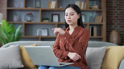Serious young asian woman works on laptop sitting on sofa in living room at home. Thoughtful female