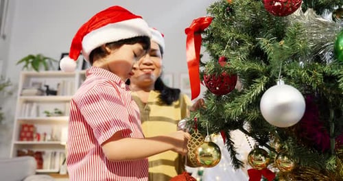 Asian boy mother wearing santa hats decorating christmas tree together hanging golden ornament with