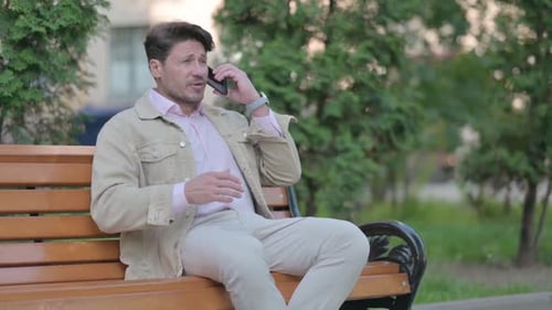 Upset Middle Aged Man Arguing on Phone while Sitting Outdoor on a Bench