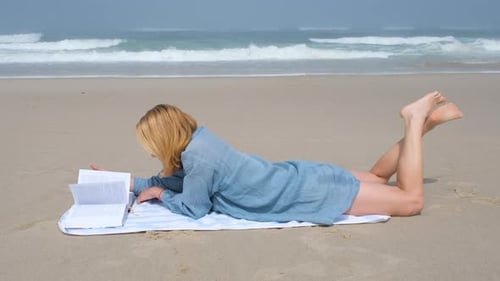A Young Woman Lies on Her Stomach on a Beach Towel Reading a Book By the Atlantic Ocean She Enjoys a