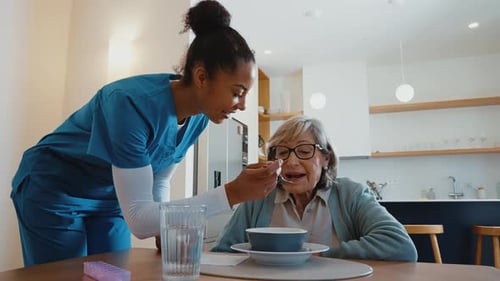 Young Woman Feeds Senior Woman Soup at Table