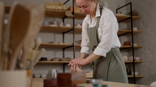 Craftswoman in Apron Joyfully Shapes Pottery Clay in a Cozy Ceramic Workshop with Shelves of Pottery