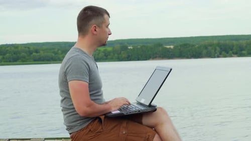 Man Typing on Laptop by Lake on Dock