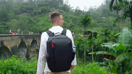 Man in Jungle Near Bridge Action Man with Backpack Looks Back at Hiking Trail in Jungle Handsome Man