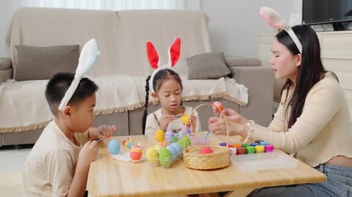 A Mother and Kids with Bunny Ears Happily Paint Easter Eggs at a Table Near a Festive Basket