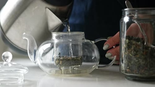 Girl Pouring Hot Water From The Kettle Into Teapot With Green Tea In The Kitchen