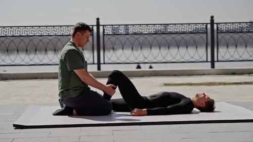 Male Physiotherapist Stretching Leg of Men Athlete on a Mat Outdoors By the River Focused