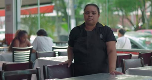 Female restaurant worker standing in dining area with neutral expression, resting hand on chair in
