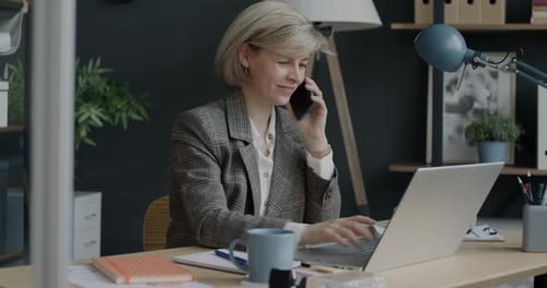 Portrait of Cheerful Businesswoman Speaking on Mobile Phone and Using Laptop Indoors in Office
