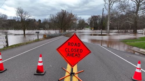 Road closed ahead sign. Aerial rising shot over roadblock revealing flooded street with brown murky