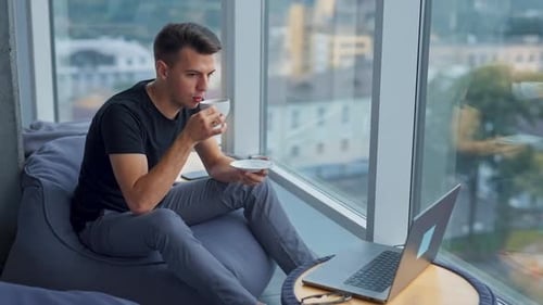 Brunetter junger Mann trinkt in der Mittagspause Kaffee und sitzt am Panoramafenster.