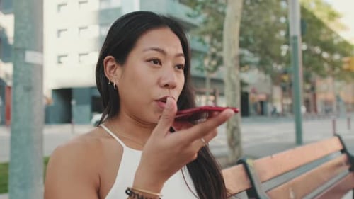 Close up, young woman sitting on bench sends voice message with mobile phone