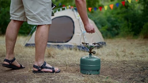 Close Up a Man in Gray Shorts Puts a Gas Burner and a Kettle on It to Heat Water in the Forest While