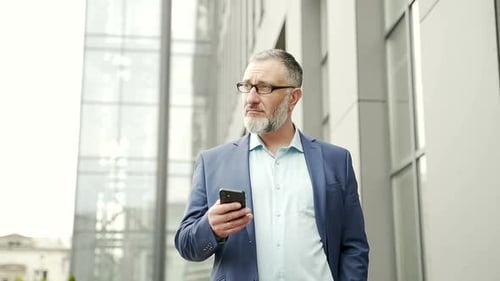 Serious thoughtful business man uses a smartphone while standing near office building outside.