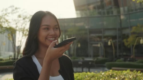 Portrait of Happy Asian Woman Records Voice Message on Smartphone in City Park