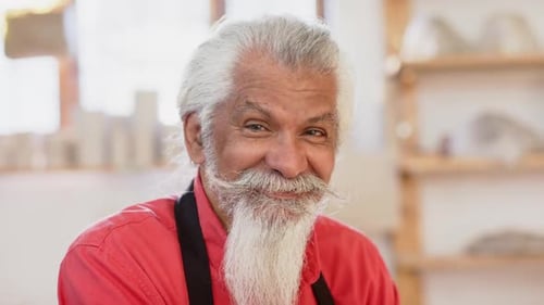 Happy potter with long beard looking on camera and smiling in pottery studio, slow motion