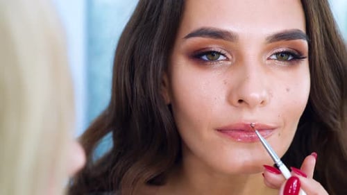 Nahaufnahme einer Visagistin, die mit einem Pinsel Lippenstift auf die Lippen einer jungen brünetten Frau aufträgt