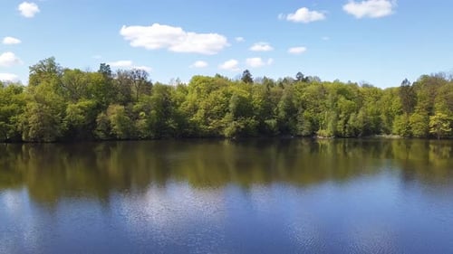 Aerial. Small lake surrounded by green trees. Blue sky, white clouds. Shot from a low flying drone.
