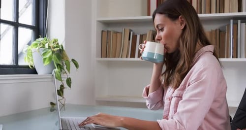 Woman Working at Laptop Drinks from Mug at Desk
