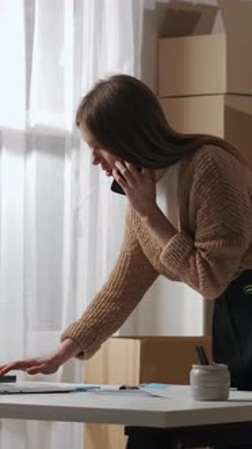 Focused Young Woman Entrepreneur Talking on Phone While Working at Desk in Home Office Surrounded By