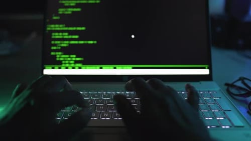 Hände tippen auf einer Laptoptastatur mit Hintergrundbeleuchtung in einem dunklen Raum mit grünem Code auf dem Bildschirm