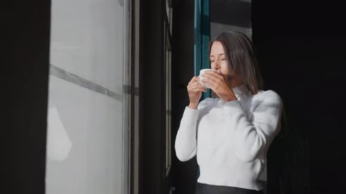 Young female stand near apartment window and enjoy morning coffee in sunlight