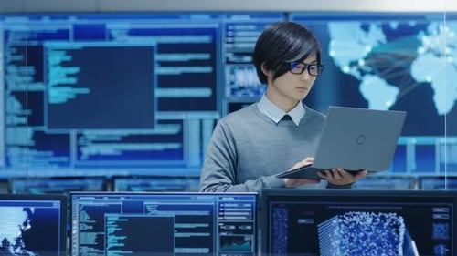 In the System Control Room IT Technician Holds and Works on a Laptop, in Background Multiple Displa