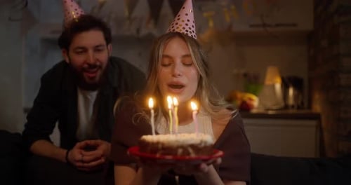 Happy Blonde Girl in a Party Cap Blowing Out Candles on a Cake While Celebrating a Birthday with Her