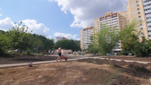 Worker with Bare Chest Pushes Cart Along Park Road in City