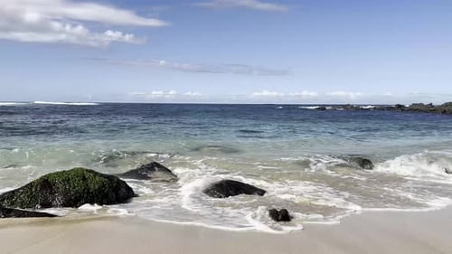 Crystal clear waves gently crash against moss-covered rocks on a pristine Hawaiian beach, capturing