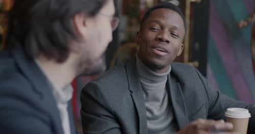 Two Young Businessmen in Suits Talking and Drinking Coffee During Informal Meeting in Cafe