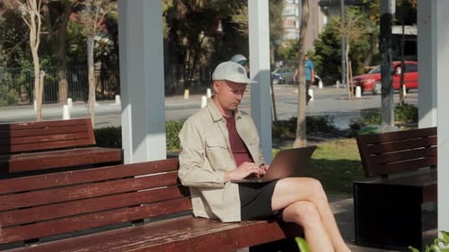 A Young Man is Sitting on a Bench in the Park and Working on a Laptop