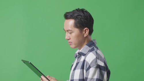 Nahaufnahme eines asiatischen Mannes mit Tablet beim Gehen auf grünem Bildschirm im Studio von der Seite