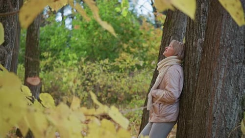 Thoughtful Woman Leans on Tree Reaching for Falling Leaf in Colorful Autumn Forest