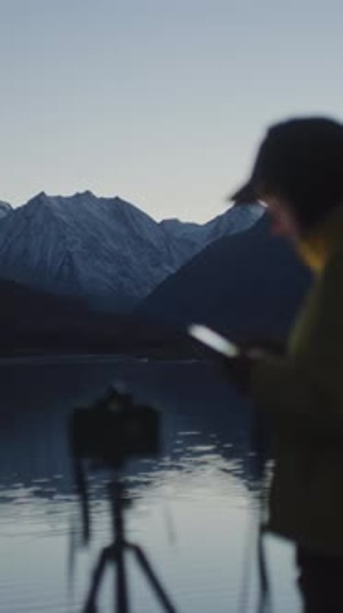 Photographer with Camera and Smartphone By Mountain Lake at Dusk