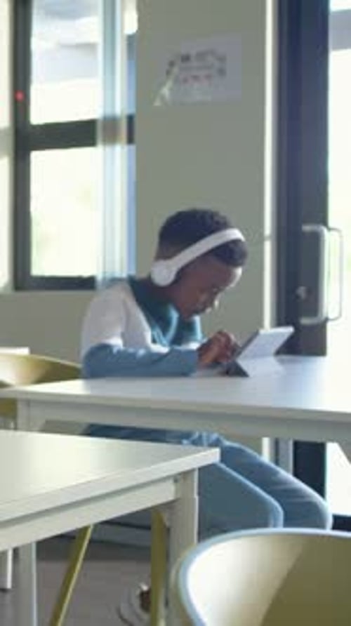 Vertical video: Tablet lighting, student tapping tablet at classroom table with headphones studying