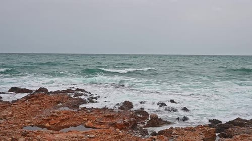 Sea in a bad weather day, gray sky, blue waves crashing into orange rocks of the coastline.