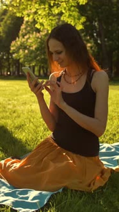 Vertical of Sexy Red Hair Girl Enjoying Sunny Day Sitting on Bench in Green Summer Park Using Mobile