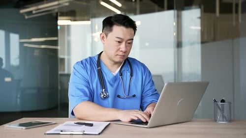 Confident asian doctor in blue coat typing on laptop sitting at workplace in hospital office.