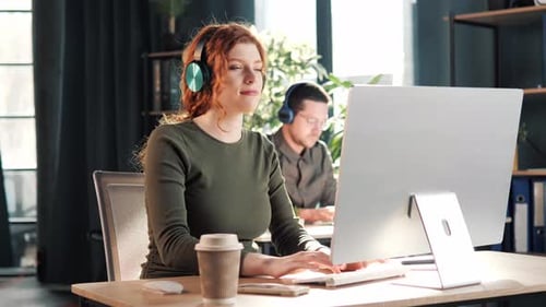 Young Woman with Red Hair Listens Music Through Headphones in Office