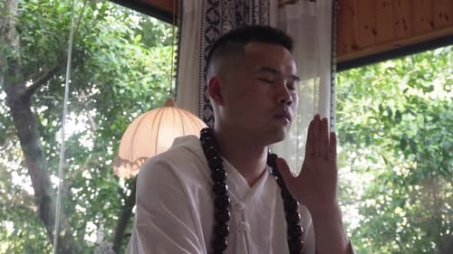 A man meditates indoors with eyes closed, wearing white robes and wooden beads, surrounded by greene