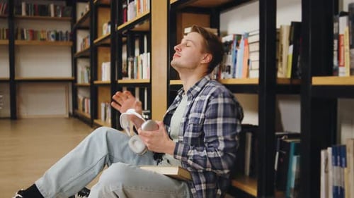 Young Tired Man Sits on the Floor of a Library Enjoying a Book While Listening to Music with