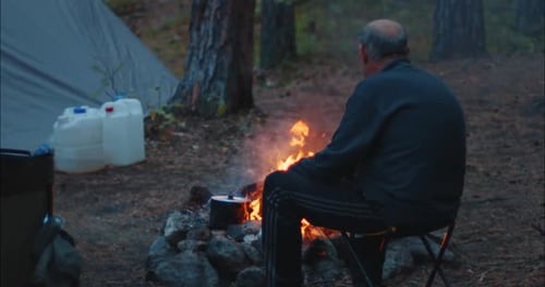 Un anciano excursionista se sienta en la silla cerca de la hoguera y la observa