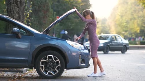 Puzzled Female Driver Standing on a City Street Near Her Car with Popped Up Hood Looking at Broken